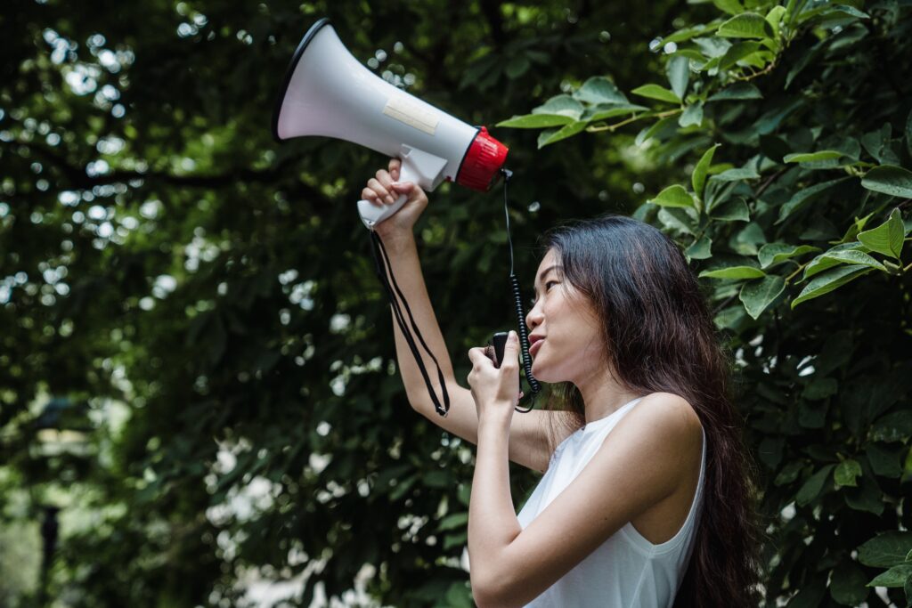 An Asian woman is using a megaphone with a large tree with green leaves in the background.